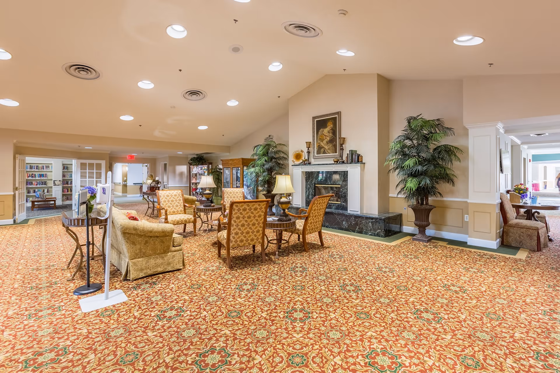 A spacious and well-lit common area in a senior living facility featuring patterned carpet, several upholstered chairs and small tables arranged around a fireplace with a painting above it. There are large potted plants, lamps on tables, and a bookshelf visible in the background. The ceiling has recessed lighting and air vents.