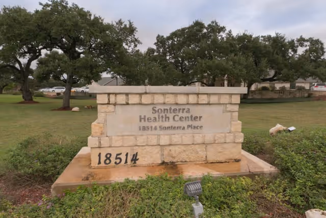 Stone sign for Sonterra Health Center with the address 18514 Sonterra Place, surrounded by grass, bushes, and trees in the background under a cloudy sky.
