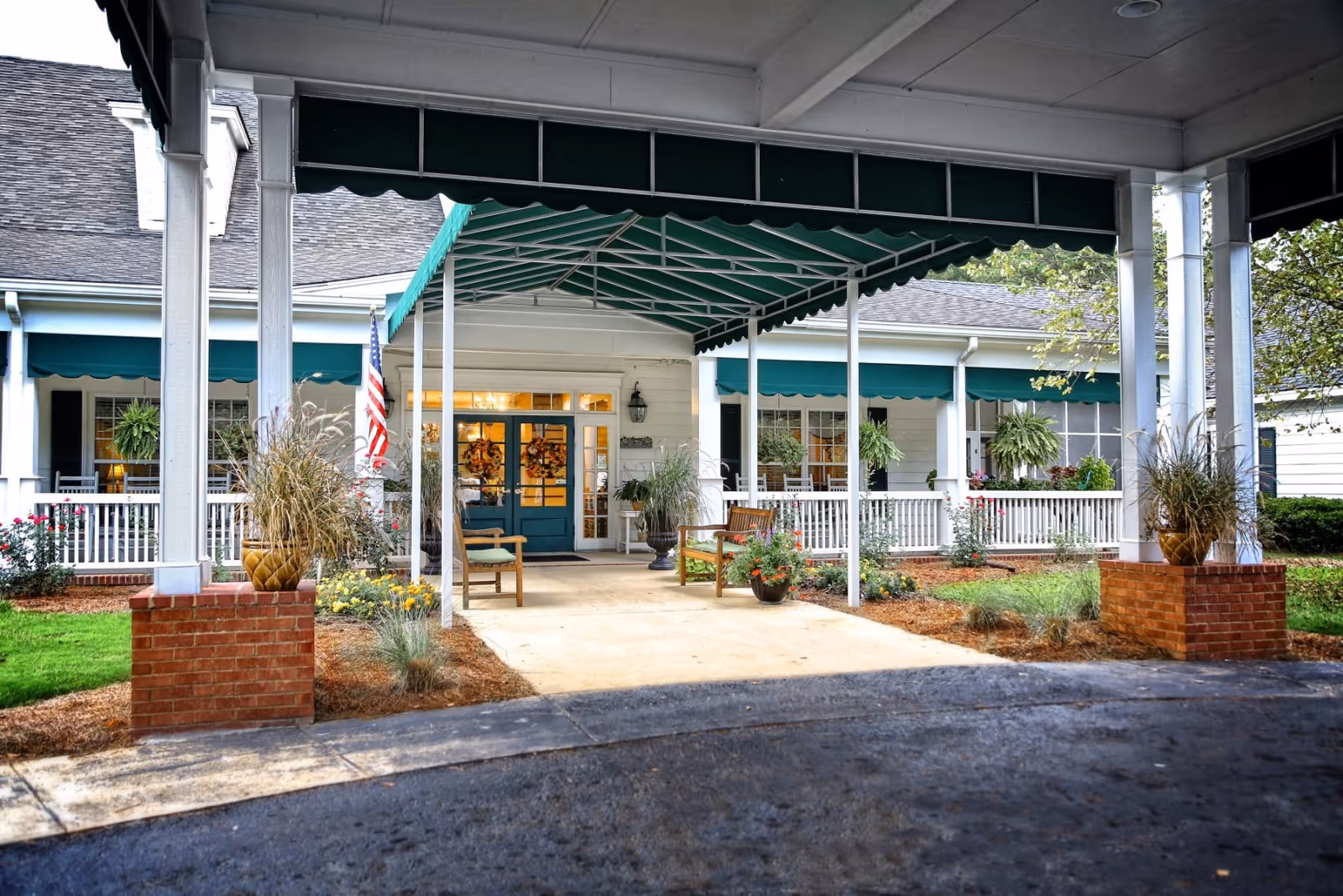 Entrance of a senior care facility with a covered driveway supported by white columns and brick bases. There are green awnings over the windows and entrance, potted plants, and two wooden benches near the doorway. An American flag is displayed near the entrance.