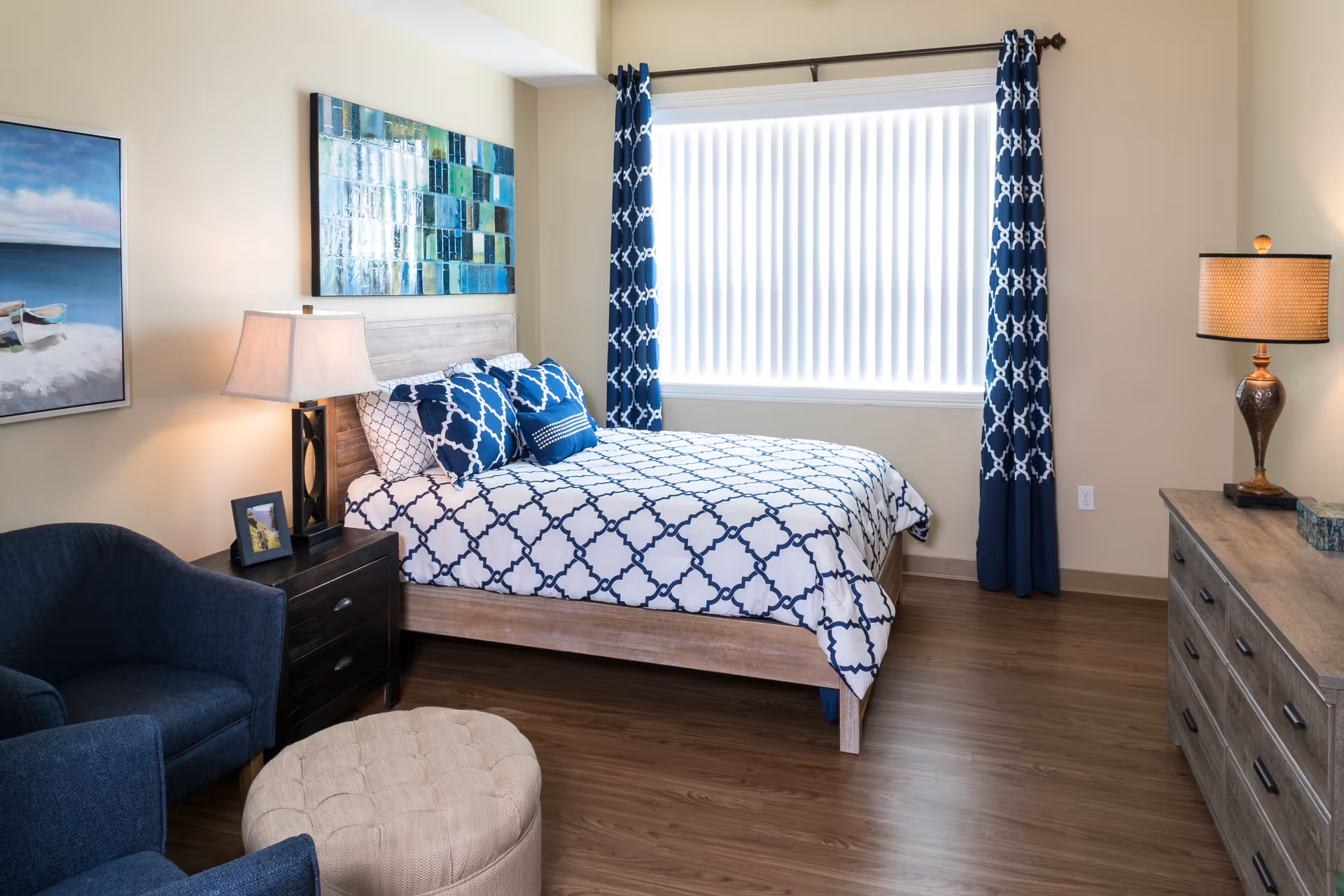 A bedroom with a wooden bed frame and a bed dressed in white and navy blue patterned bedding. There are matching navy blue curtains on the window with vertical blinds. The room also features a dark wooden nightstand with a lamp and a framed photo, a blue upholstered armchair, a beige tufted ottoman, a wooden dresser with a lamp, and two paintings on the walls.