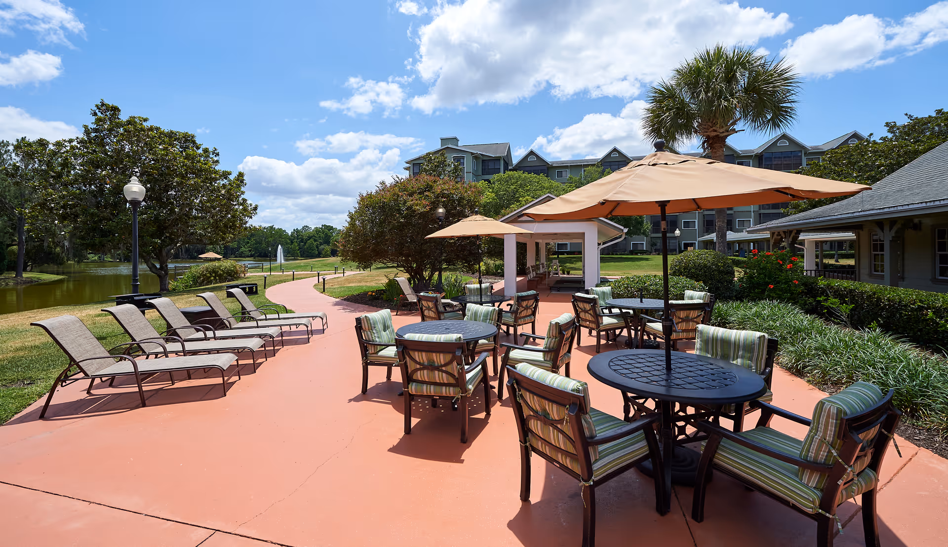 Sunlit outdoor patio area with umbrella-covered tables and cushioned chairs, lounge chairs along a walkway, a pond, and a multi-story building in the background.
