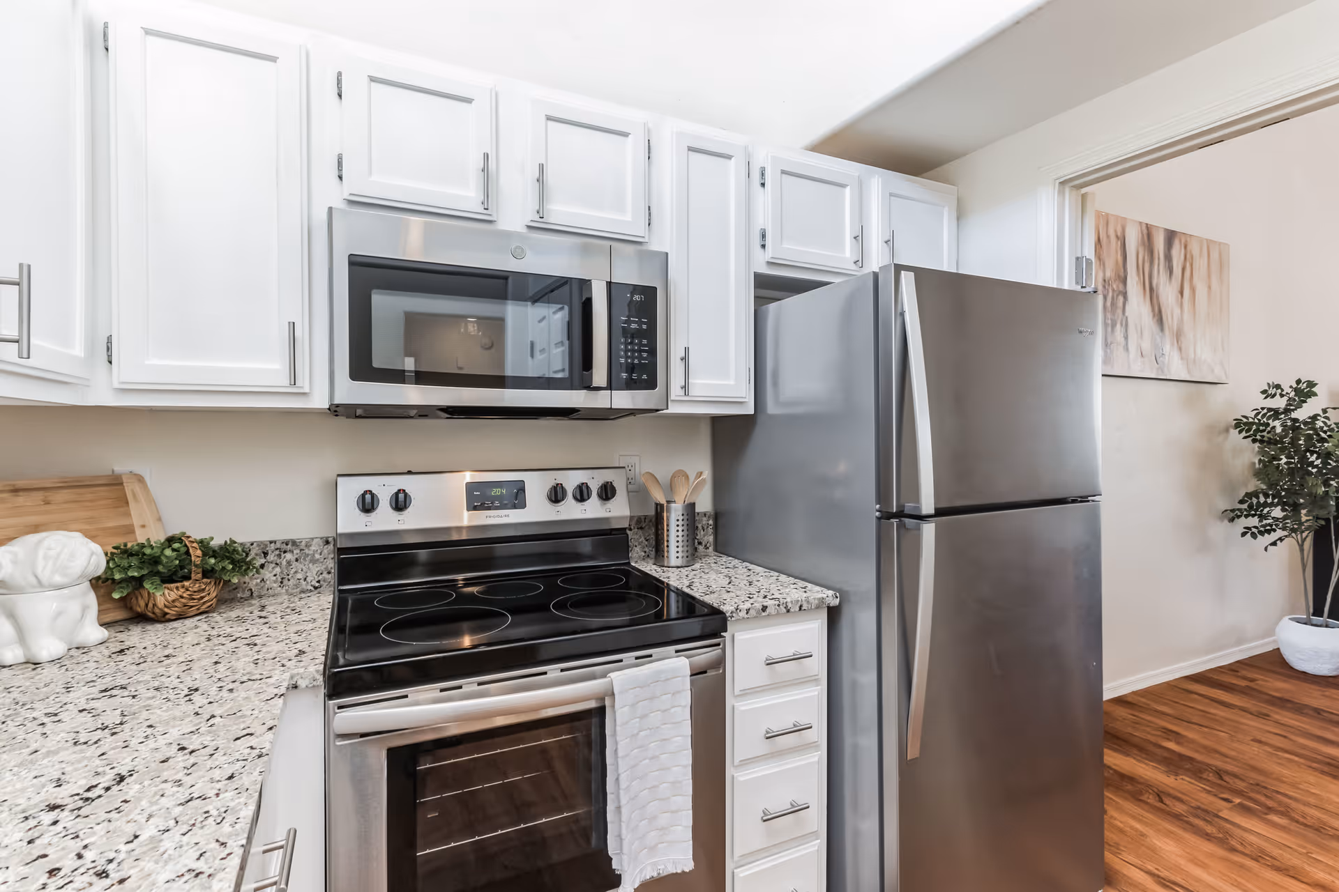 Modern kitchen with white cabinets, a stainless steel microwave above a black and stainless steel electric stove, a stainless steel refrigerator, granite countertops, and wooden flooring. A small plant and kitchen utensils are visible on the countertop.