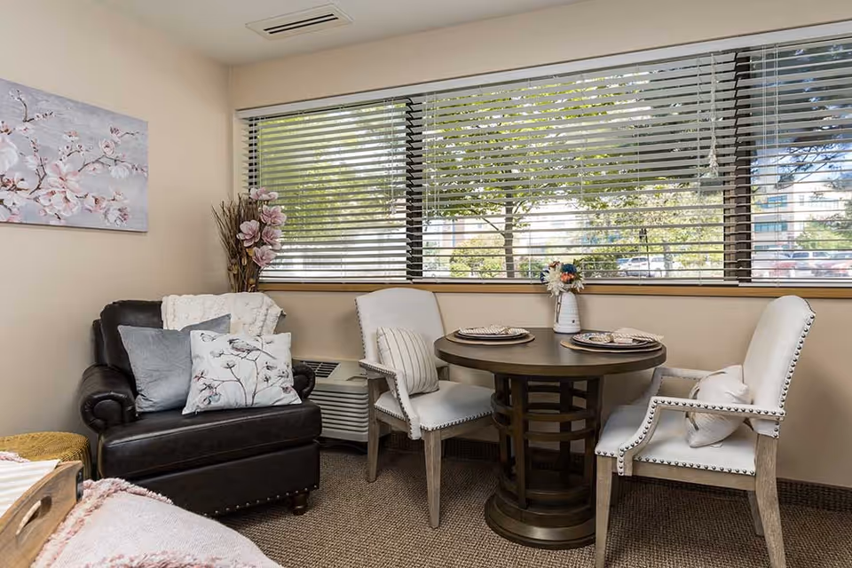 Cozy sitting area with a leather armchair, round wooden dining table and three upholstered chairs set by a large window with blinds.