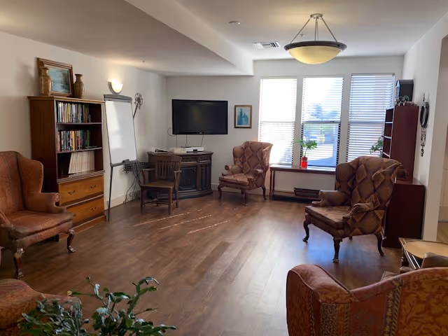 A cozy living room with wooden flooring, featuring four upholstered armchairs arranged around the room. There is a wooden bookshelf filled with books, a flat-screen TV mounted on the wall, a small wooden table with a chair, and a whiteboard on a stand. Large windows with blinds allow natural light to fill the space, and a ceiling light fixture hangs overhead. Some potted plants are placed near the windows.