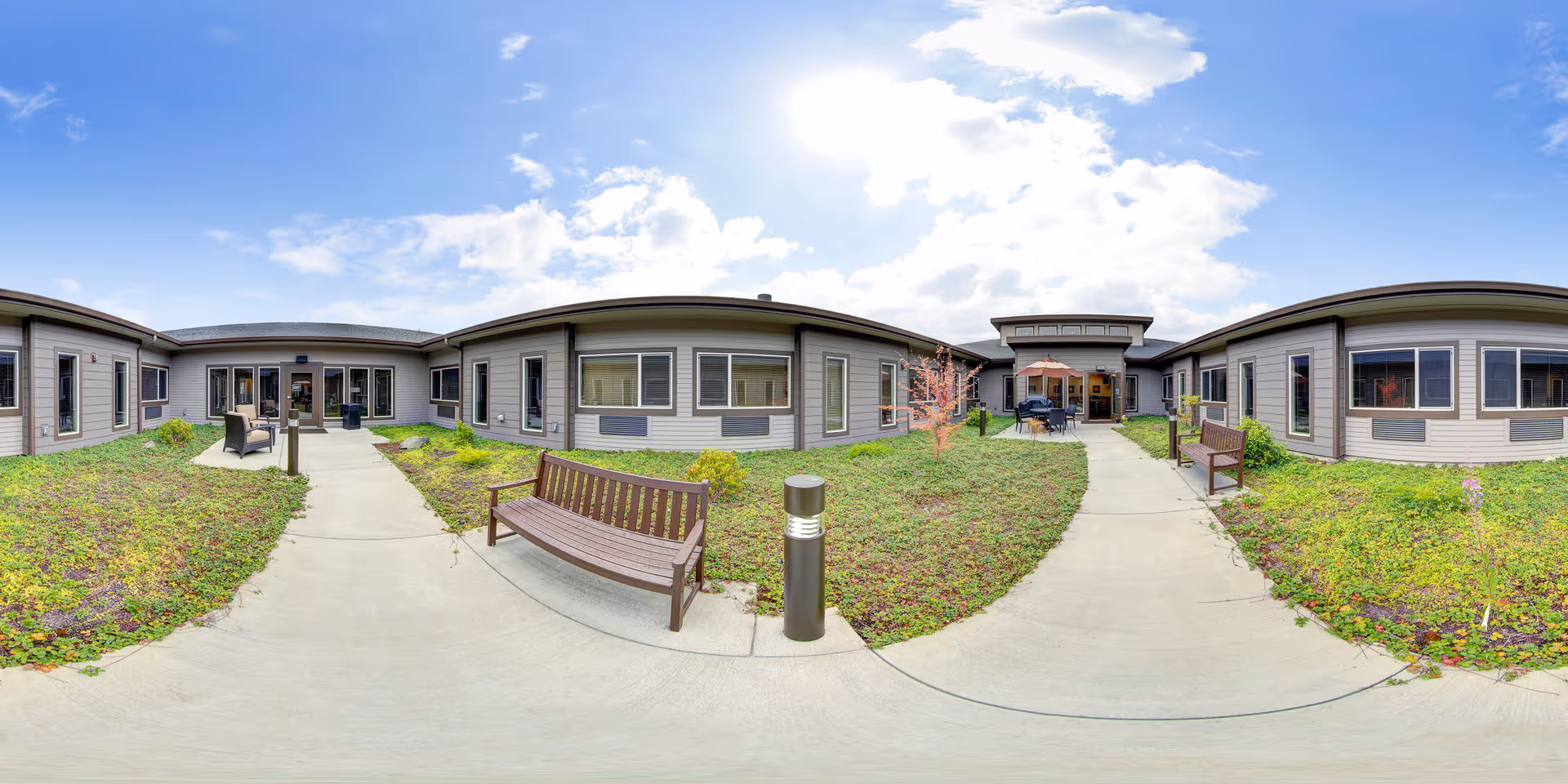 Outdoor courtyard area of Avamere Transitional Care of Puget Sound with benches, a concrete walkway, green plants, and a partly cloudy sky above. The courtyard is surrounded by single-story building sections with windows and doors, and there is outdoor seating with an umbrella near the entrance.