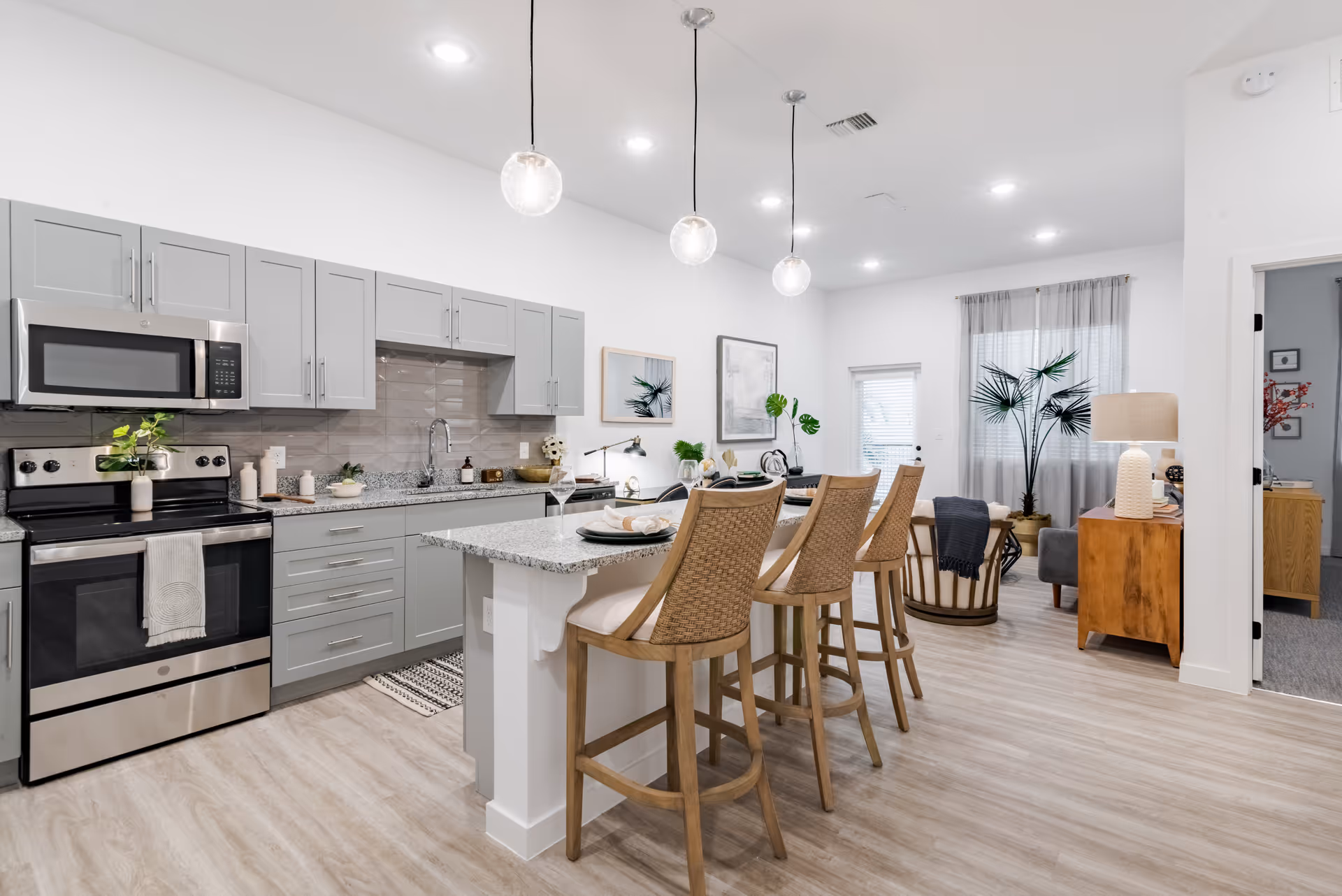 Modern open-concept kitchen and living area with light wood flooring, gray cabinets, stainless steel appliances, a granite countertop island with three wooden bar stools, pendant lights hanging above, and a cozy living space with plants and a wooden side table with a lamp.