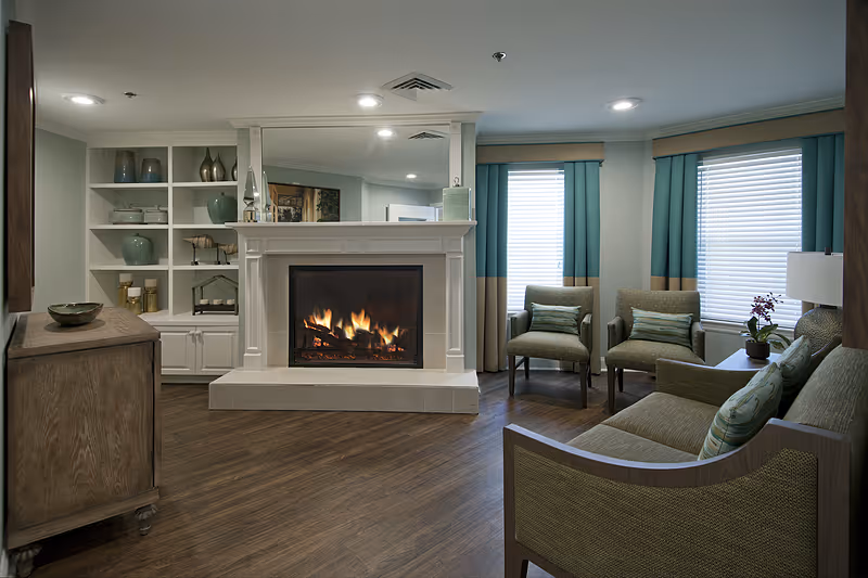 A cozy living room with a lit fireplace in the center, flanked by built-in white shelves with decorative items. The room has wooden flooring, a wooden cabinet on the left, and seating including a sofa and two armchairs with green cushions near two windows with teal and beige curtains. A small table with a lamp and a potted plant is beside the sofa.