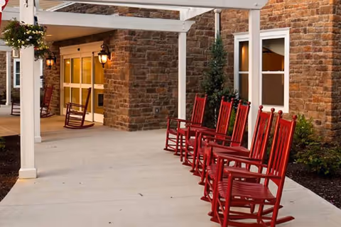 Covered front entrance of a brick building with red rocking chairs lined up on a concrete porch.