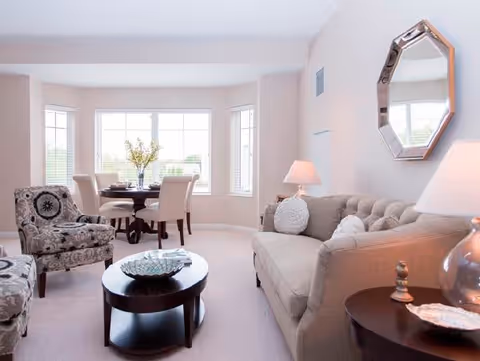 A bright living room with a beige tufted sofa, two patterned armchairs, a round dark wood coffee table with a decorative bowl, and a small round side table with a lamp. In the background, there is a dining area with a round table and four upholstered chairs near large windows letting in natural light. A decorative mirror hangs on the wall above the sofa.