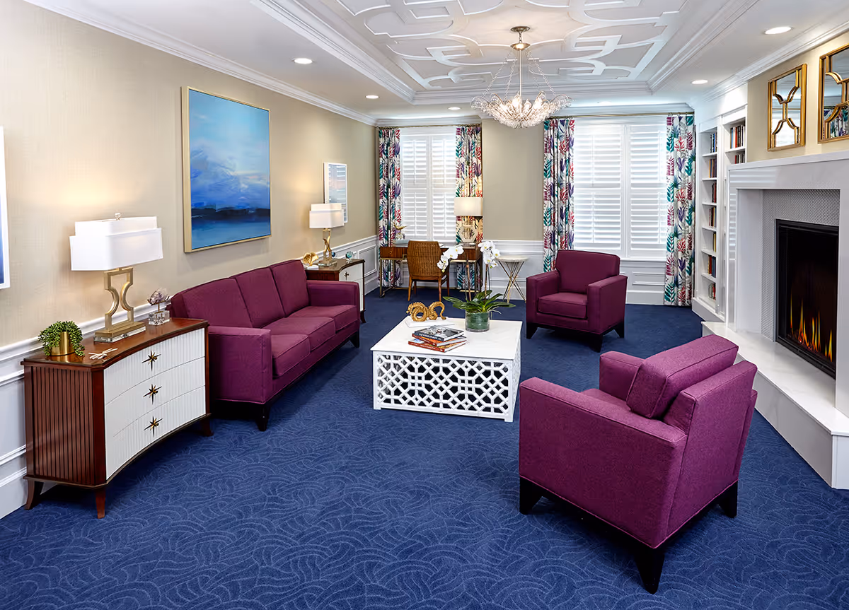 A bright and elegant living room with purple upholstered sofa and armchairs arranged around a white decorative coffee table. The room features a blue carpet, a modern fireplace, colorful floral curtains, a wooden sideboard with lamps, and a ceiling chandelier. There are also bookshelves and framed artwork on the walls.