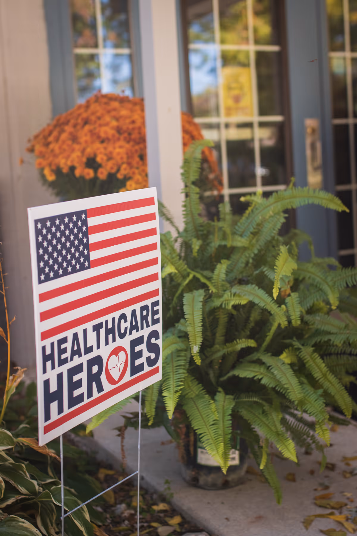 A 'Healthcare Heroes' yard sign with an American flag displayed among potted plants in front of a building entrance.