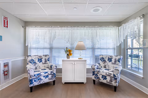A bright sitting area with two armchairs upholstered in white fabric with blue floral patterns, positioned on either side of a small white cabinet. On top of the cabinet is a table lamp and a small vase with yellow flowers. The background features large windows with white blinds and lace curtains, allowing natural light to fill the room. The floor is light wood, and the walls are painted in a soft neutral color.