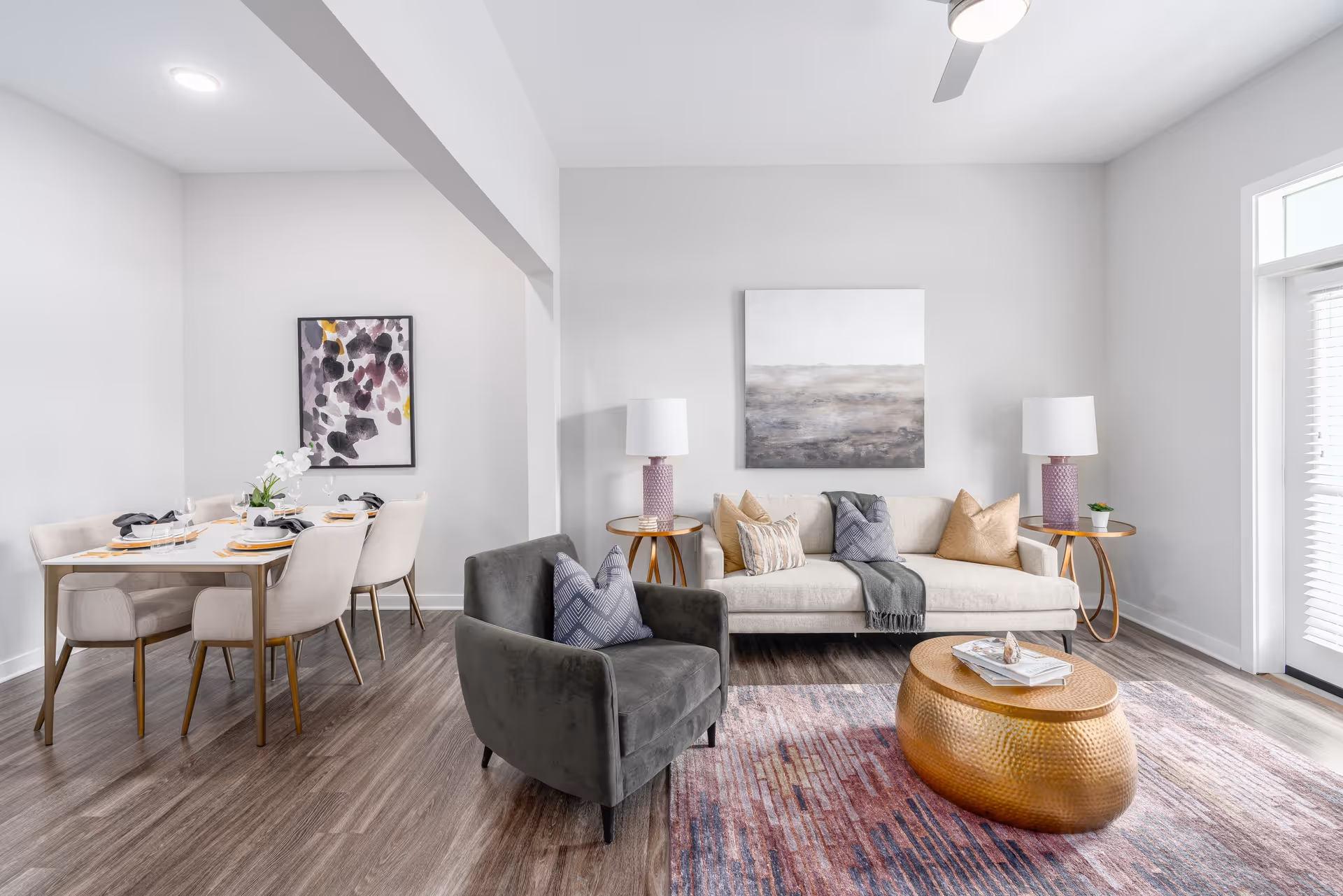 A modern living room and dining area featuring a beige sofa with decorative pillows, a dark gray armchair with a patterned pillow, a gold oval coffee table on a multicolored rug, and two side tables with purple lamps. The dining area has a white table set with plates, glasses, and napkins, surrounded by six beige upholstered chairs. The walls are light gray with two abstract paintings, and there is a ceiling fan and large windows with blinds allowing natural light in.