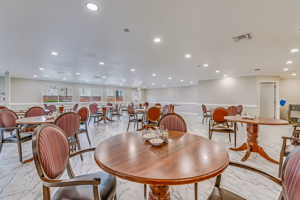 A spacious dining room with multiple round wooden tables and cushioned chairs with striped upholstery. The room has a bright, clean atmosphere with white walls, large windows, and a marble-patterned floor. The ceiling is equipped with numerous recessed lights.