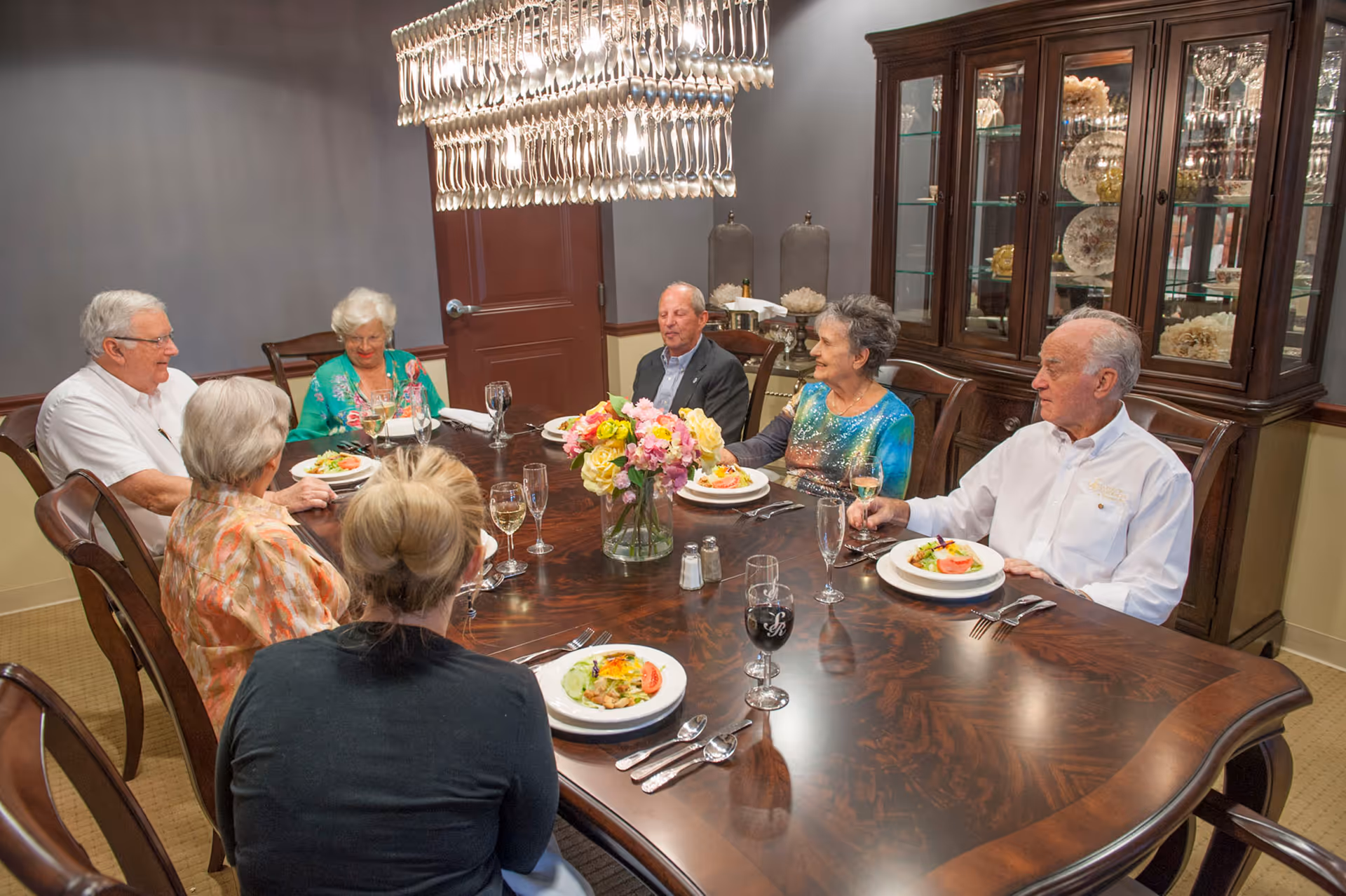 Seven older adults seated around a formal dining table with plated salads, wine glasses, and a floral centerpiece in a decorated dining room.