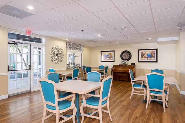 A bright dining room with several square tables surrounded by teal cushioned chairs with light wood frames. The room has light yellow walls, wood flooring, and a white ceiling with recessed lighting. There is a piano against the far wall with a floral arrangement on top, flanked by two framed pictures and a decorative clock. A glass door and window on the left side let in natural light.