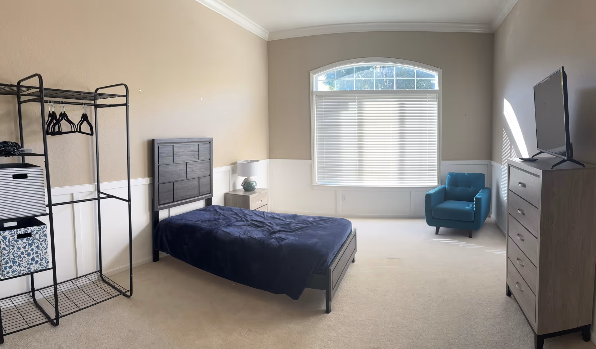 A bright bedroom with beige walls and white wainscoting. The room features a single bed with a dark wooden headboard and a navy blue blanket, a small nightstand with a lamp, a metal open wardrobe rack with hangers and storage boxes, a blue armchair near a large window with white blinds, and a wooden dresser with a flat-screen TV on top.