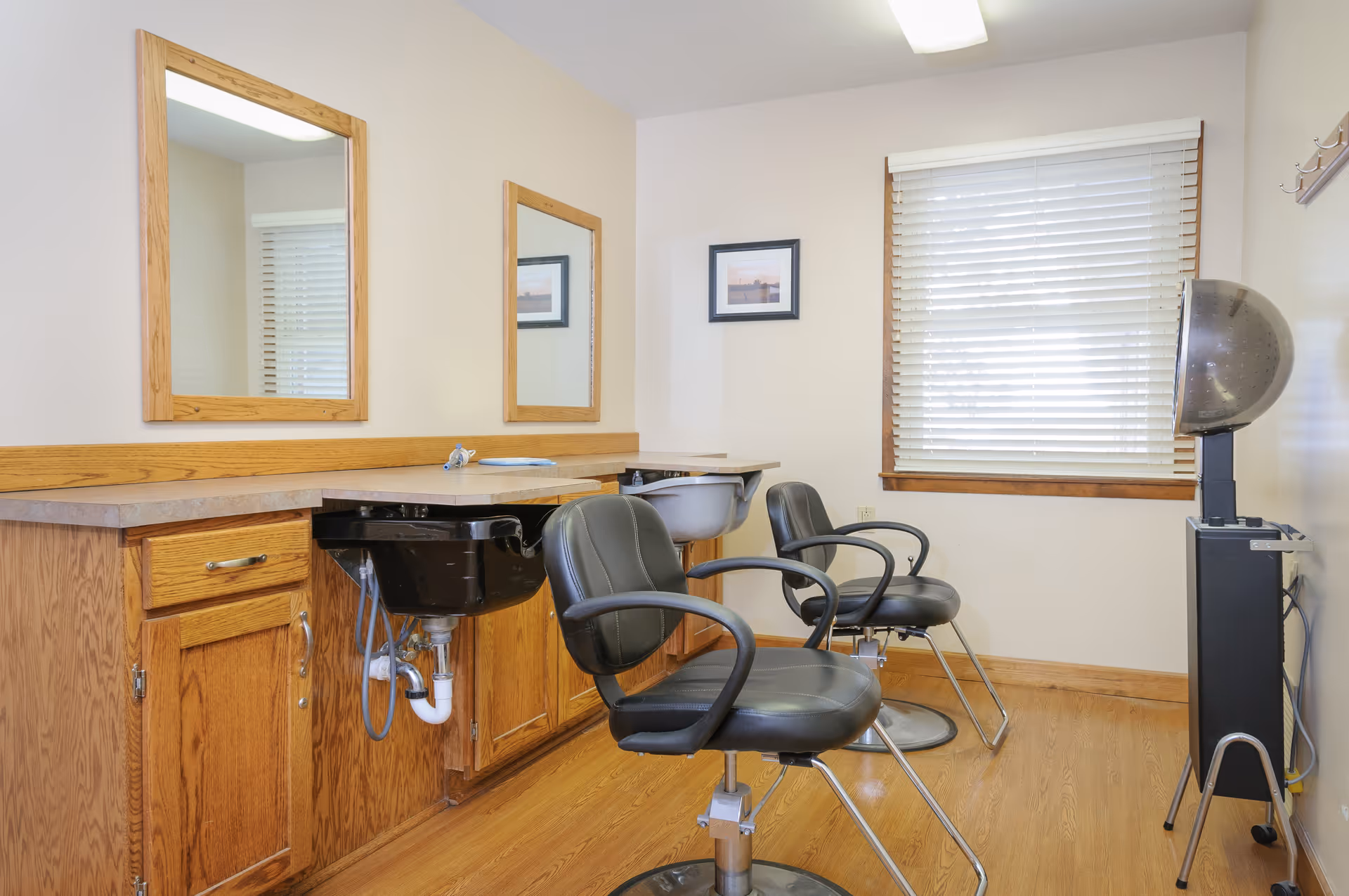 Interior view of a small salon area in a senior living facility with two black salon chairs, two mirrors mounted on the wall above a countertop with wooden cabinets, a black hair washing sink, a window with white blinds, and a hair dryer station.