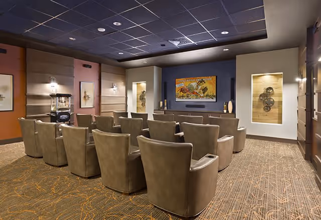A cozy theater room with three rows of comfortable leather recliner chairs facing a large screen mounted on a dark blue accent wall. The room has warm lighting, decorative wall niches with art pieces, and a patterned carpet.