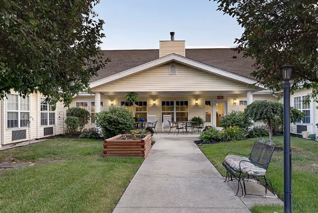Outdoor courtyard area of a senior living facility with a paved walkway leading to a covered patio. The patio has tables and chairs, surrounded by well-maintained grass, bushes, and trees. A bench and a lamp post are visible along the walkway.
