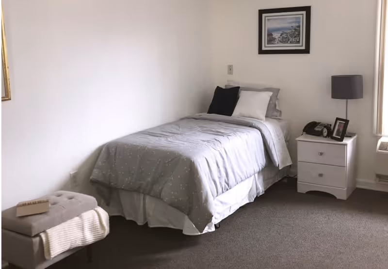Small bedroom with a single bed dressed in gray bedding, a white nightstand with a lamp and phone, a framed picture on the wall, and a small ottoman.