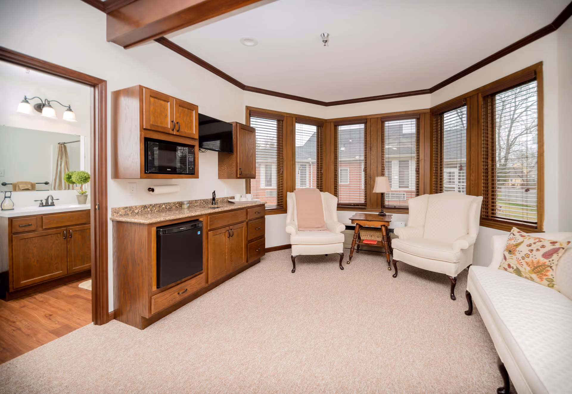 A cozy living area with beige carpet, two white upholstered armchairs, a white upholstered bench with a floral pillow, and a wooden side table with a lamp. The room has large windows with wooden blinds and wood trim. To the left, there is a kitchenette with wooden cabinets, a microwave, a small refrigerator, and a granite countertop. A doorway leads to a bathroom with wooden cabinets, a sink, and a mirror with lights above it.
