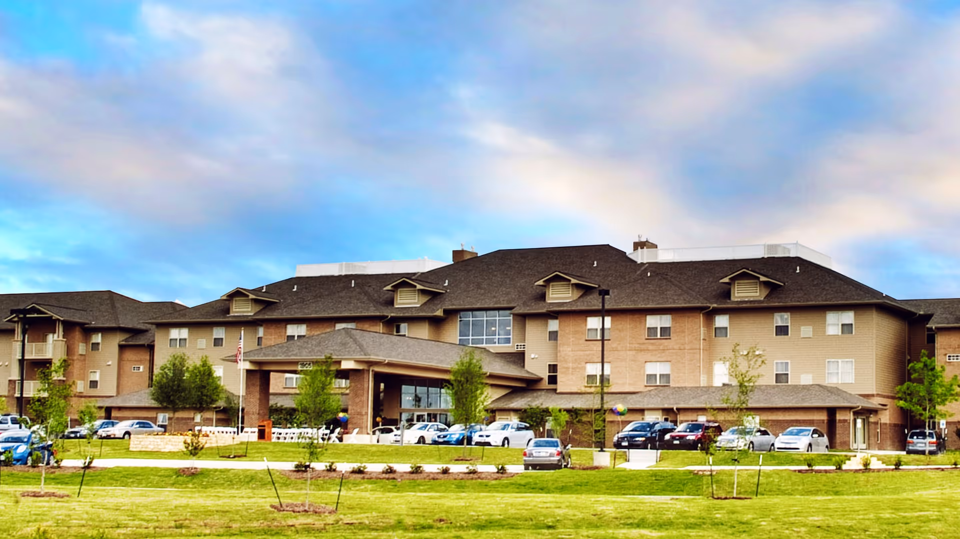 Exterior view of Lakeline Oaks Retirement Community, a large multi-story building with a pitched roof, multiple windows, and a covered entrance. The foreground features a well-maintained lawn with young trees and a parking area with several cars. The sky is partly cloudy with a blue and light pink hue.
