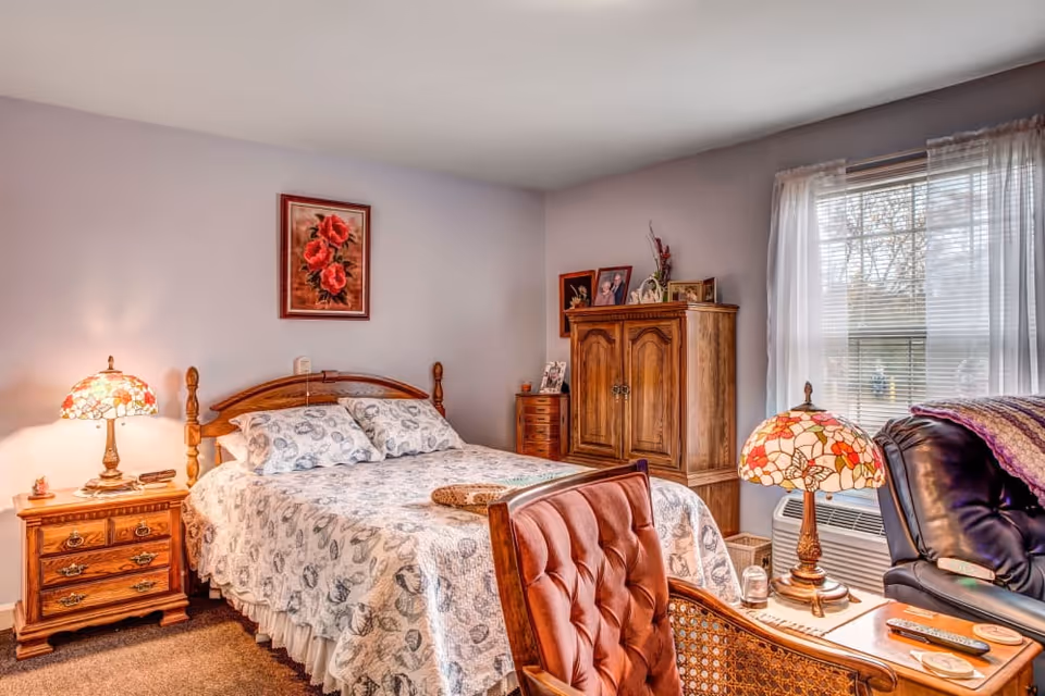 Cozy bedroom with a wooden bed and patterned bedspread, matching nightstands with Tiffany-style lamps, an armoire, and a window with sheer curtains.