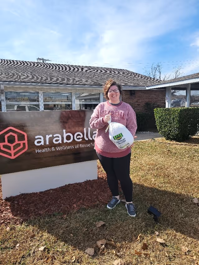 A woman stands outdoors in front of the Arabella Health & Wellness of Russellville sign holding a packaged turkey.