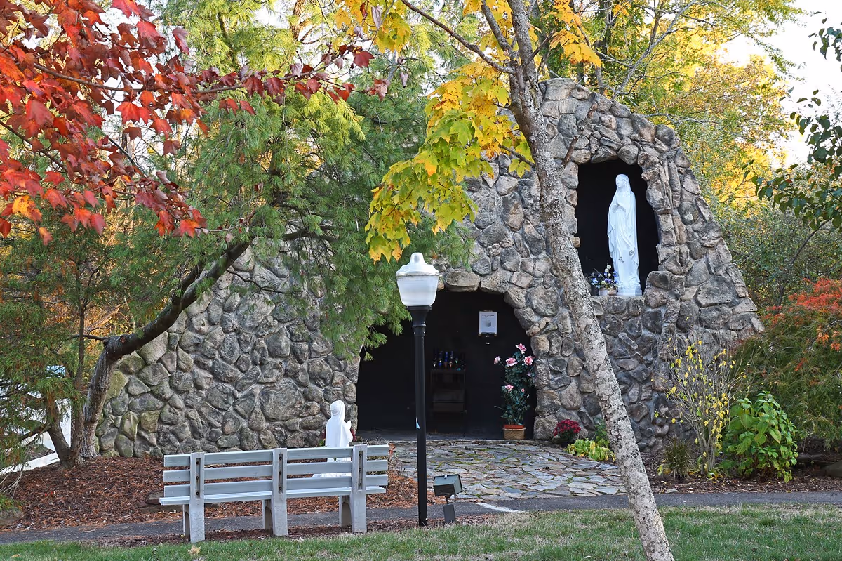 Stone grotto with religious statues, a bench and lamp post surrounded by autumn trees and landscaping.
