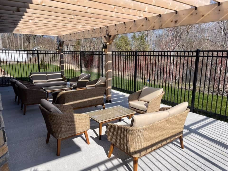 Outdoor patio area with wicker furniture including sofas and armchairs arranged around glass-top coffee tables under a wooden pergola. The space is enclosed by a black metal fence with grass and trees visible beyond.