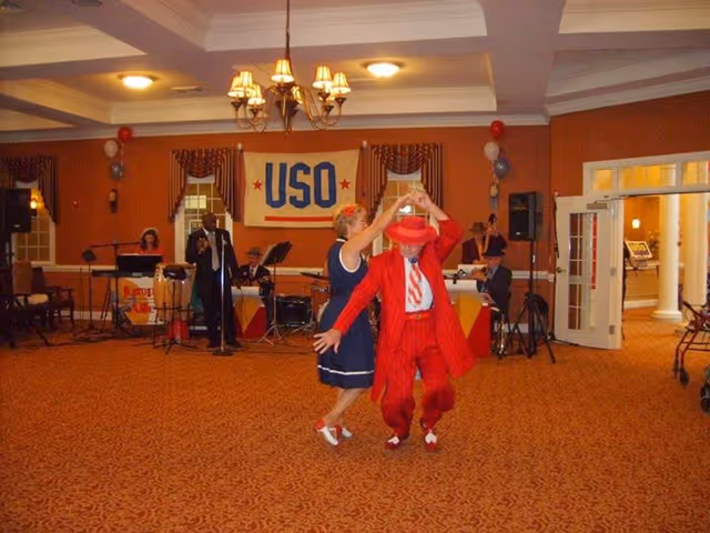 A couple dances in a carpeted activity room in front of a live band and a large 'USO' banner.