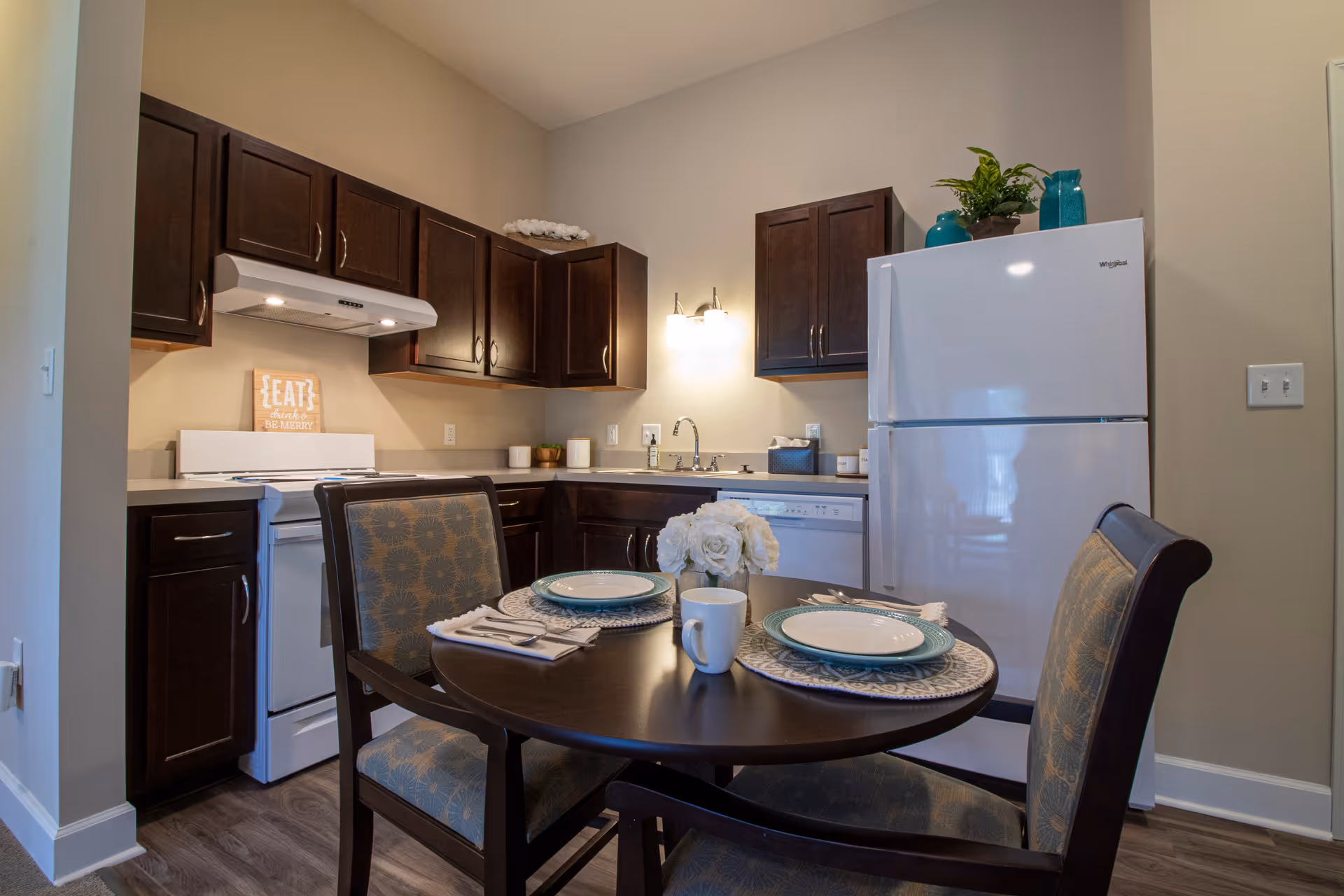 A small kitchen and dining area with dark wood cabinets, a white refrigerator, stove, and dishwasher. A round dining table is set with two place settings, including plates, napkins, and a coffee mug, with a small floral centerpiece. The kitchen has a light fixture above the sink and decorative items on top of the cabinets.