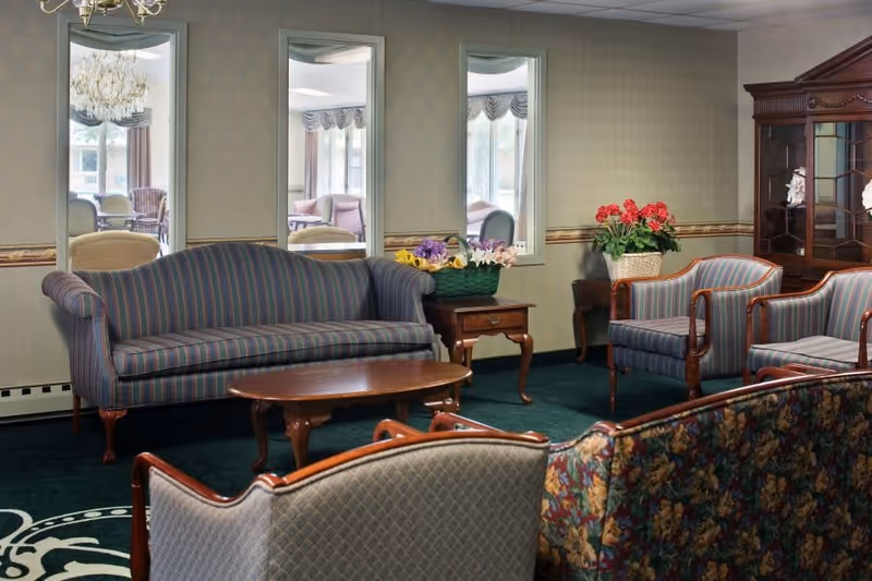 A cozy living room area in a senior living facility featuring a striped sofa, two matching armchairs, a wooden coffee table, and side tables with flower arrangements. The room has three tall mirrors on the wall, a chandelier, and a wooden display cabinet. The carpet is dark green with a decorative pattern.