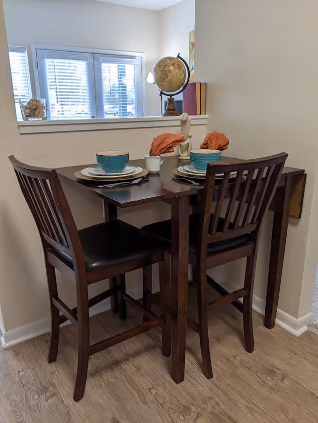 A small dining area with a dark wooden table set for two, featuring teal bowls, plates, silverware, and orange cloth napkins. Two matching wooden chairs with black cushions are positioned at the table. In the background, there is a window with white blinds, a globe, some books, and a decorative sculpture on a ledge.