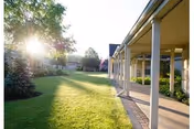 Sunlit grassy courtyard with a covered columned walkway along the side of a senior living building and trees in the background.