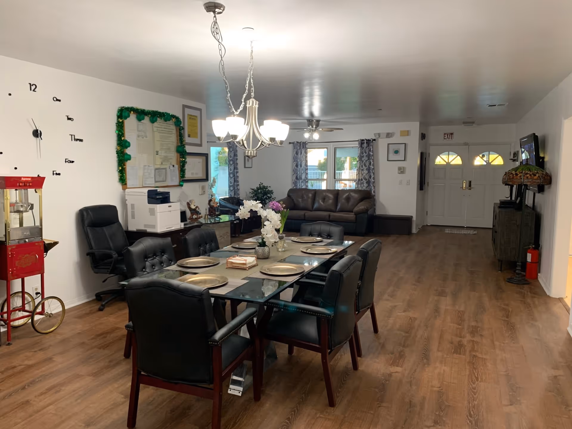 Interior view of a senior living facility room with a glass dining table set with six black cushioned chairs and place settings. A chandelier hangs above the table. In the background, there is a brown leather sofa, windows with patterned curtains, a ceiling fan, and a double door exit. On the left side, there is a red popcorn machine, a black office chair, a printer on a cabinet, and a wall clock with numbers and words. The floor is wood, and the walls are white.