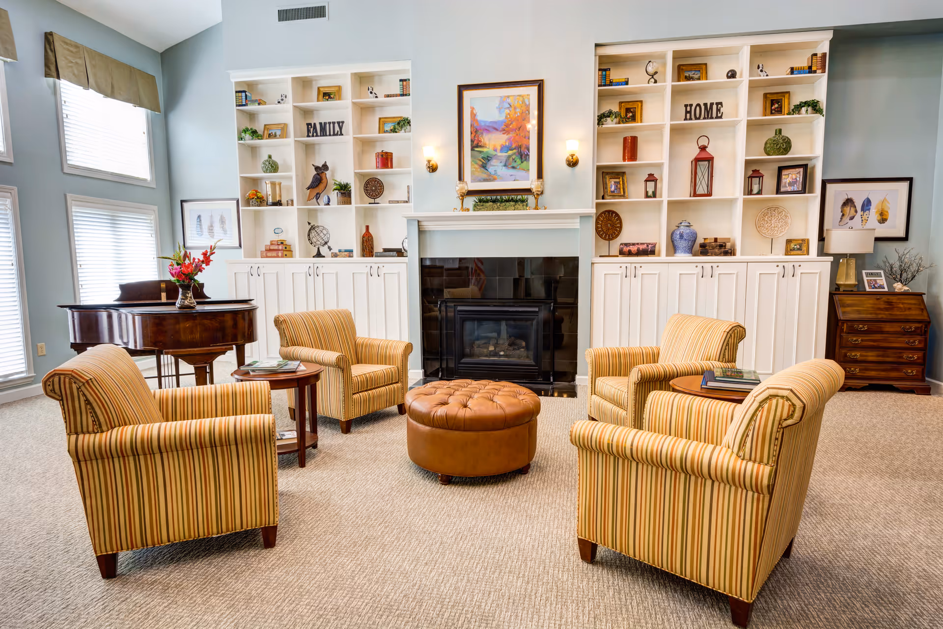 A cozy living room with four striped armchairs arranged around a round leather ottoman in front of a fireplace. Behind the fireplace are built-in white shelves decorated with books, plants, framed pictures, and decorative items. A grand piano with a vase of flowers is positioned near large windows with blinds. The walls are painted light blue, and the room has a warm, inviting atmosphere.