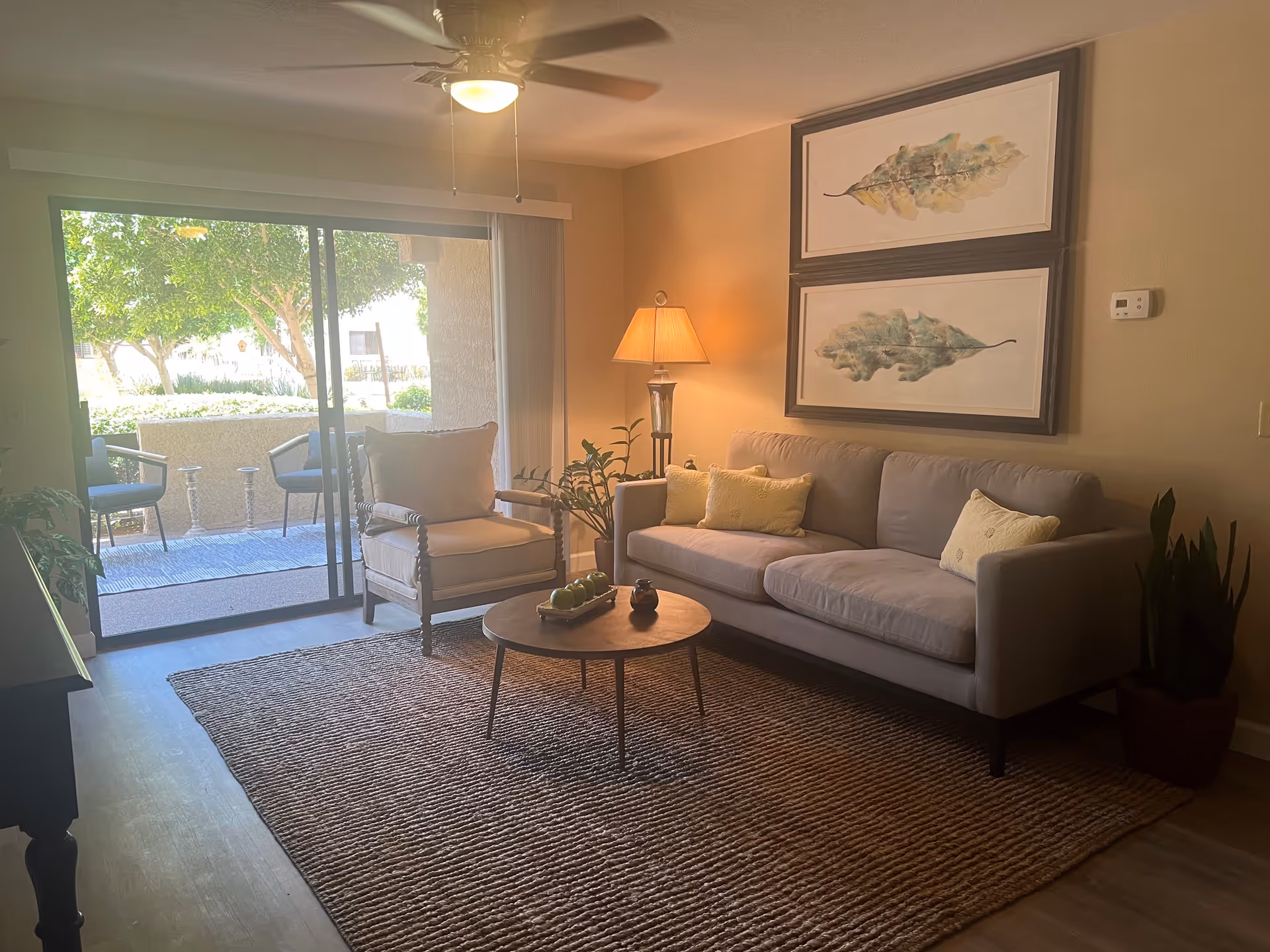 A cozy living room with a beige sofa adorned with three yellow pillows, a cushioned armchair, a round wooden coffee table with decorative items, a floor lamp, two framed leaf artworks on the wall, and a sliding glass door leading to an outdoor patio with chairs and greenery.