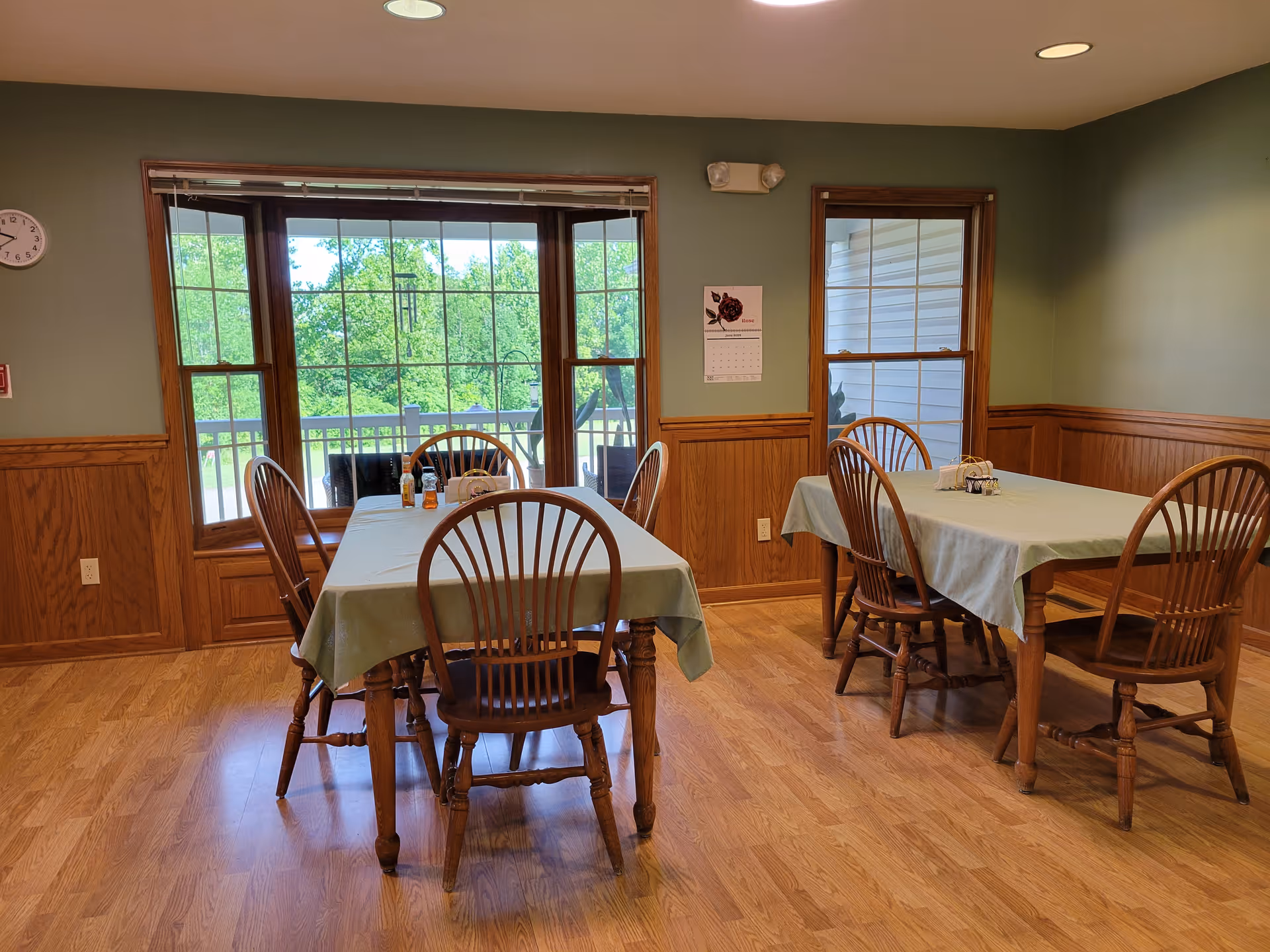 A dining room with two wooden tables covered with light green tablecloths, each surrounded by wooden chairs. Large windows and a glass door provide a view of green trees outside. The walls are painted green with wooden wainscoting, and the floor is wooden. A clock and a calendar are visible on the walls.