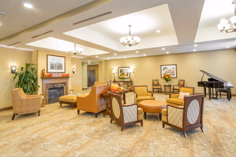 A spacious and well-lit senior living facility lounge area with beige walls and carpet. The room features multiple comfortable chairs and sofas arranged around a central round ottoman. There is a fireplace decorated with autumn-themed items and a framed picture above it. A grand piano is positioned in the corner, and the ceiling has recessed lighting and chandeliers.