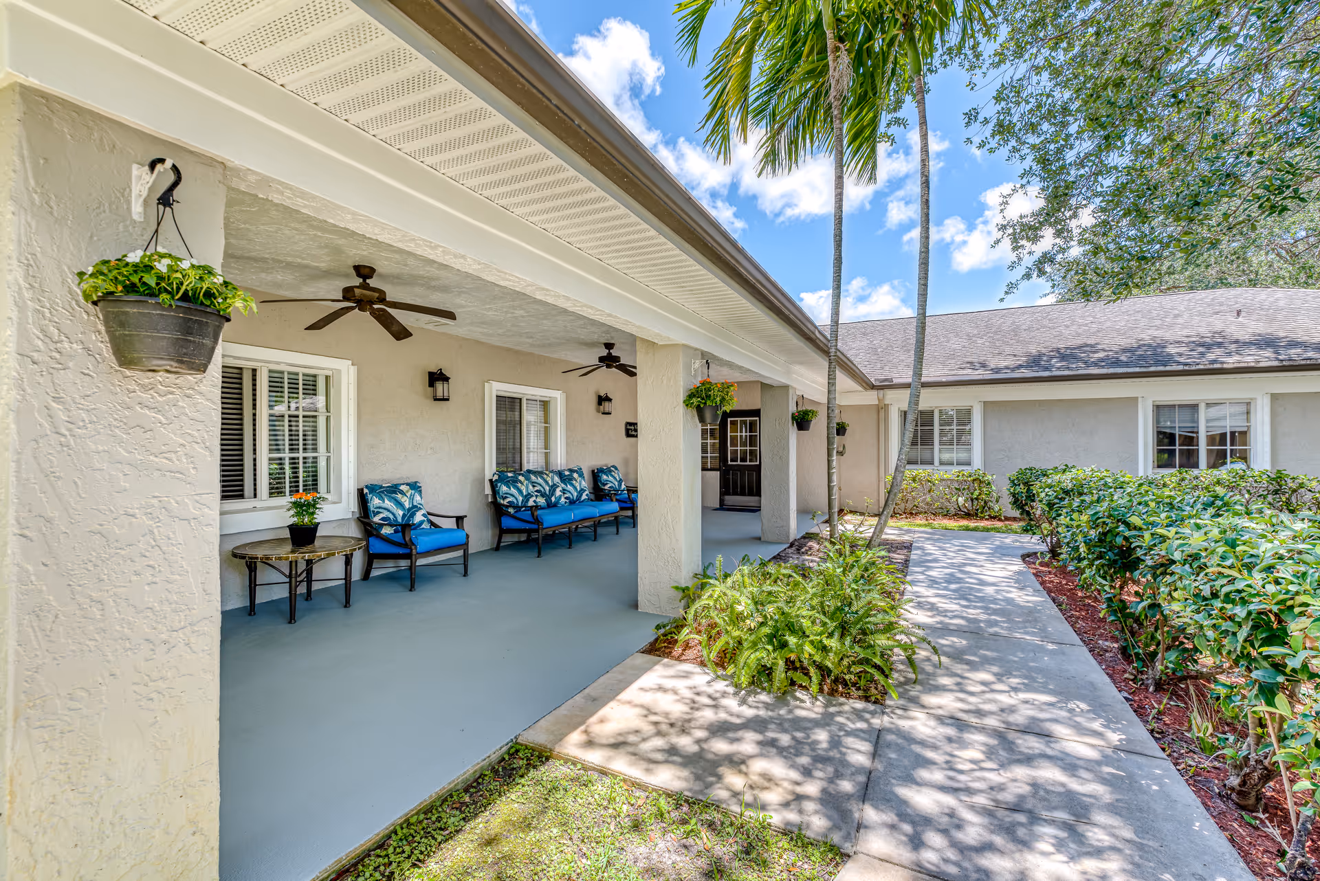 Outdoor covered patio area at a senior living facility with cushioned chairs and a small table, ceiling fans, hanging plants, and a walkway surrounded by greenery and palm trees under a blue sky with clouds.