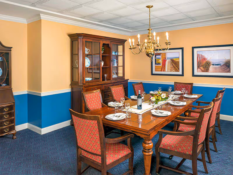 A formal dining room with a wooden dining table set for six people with plates, glasses, and silverware. The chairs have red patterned upholstery. A wooden china cabinet with glass doors displays decorative plates and glassware. Two framed beach-themed paintings hang on the yellow and blue walls. A brass chandelier hangs above the table.