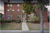 Front exterior view of a three-story brick building with multiple windows, a central entrance with steps leading up to a door, surrounded by trimmed bushes and trees on a grassy lawn.