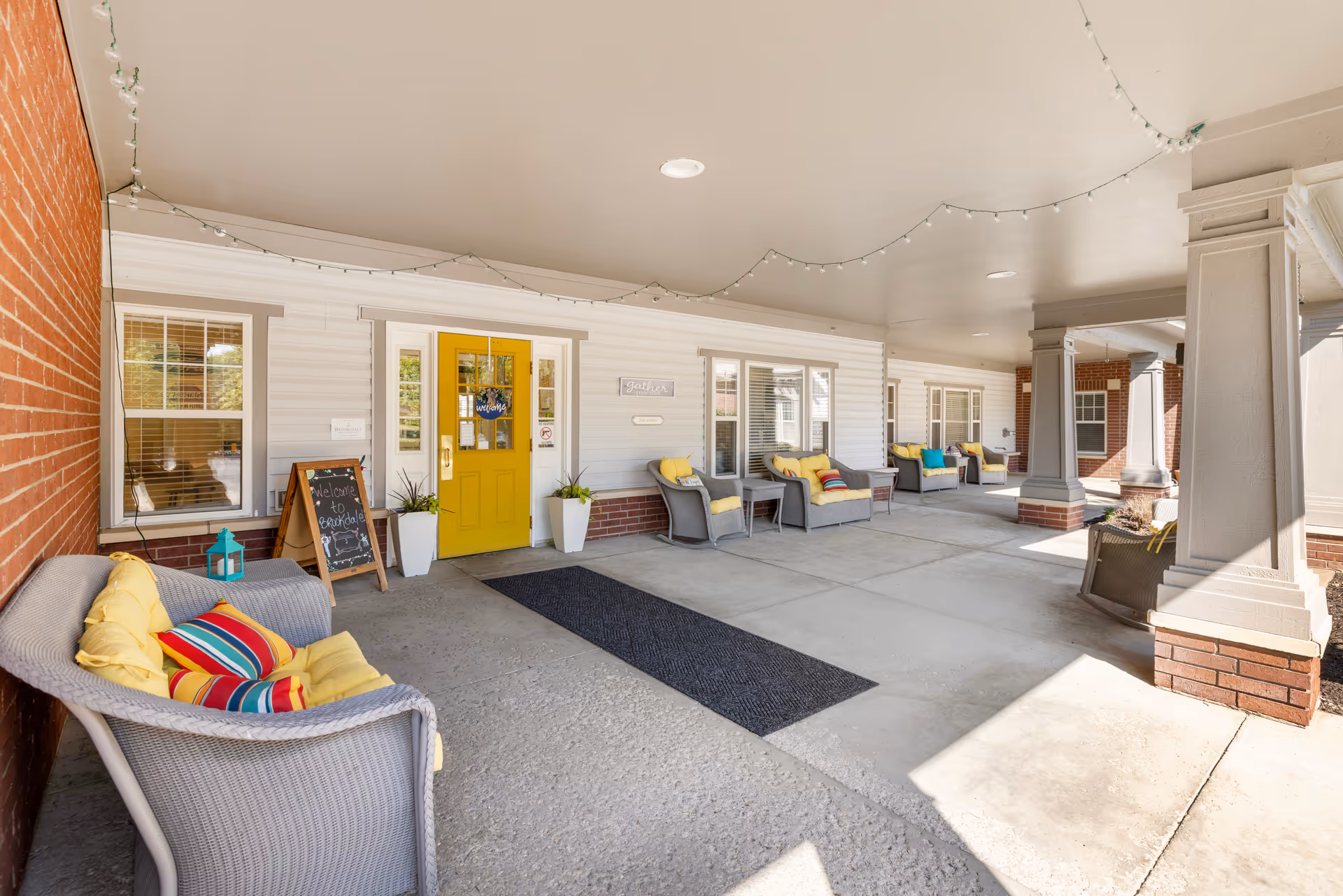 Covered entrance porch of a senior living building with a bright yellow front door, wicker seating with colorful cushions, and string lights overhead.