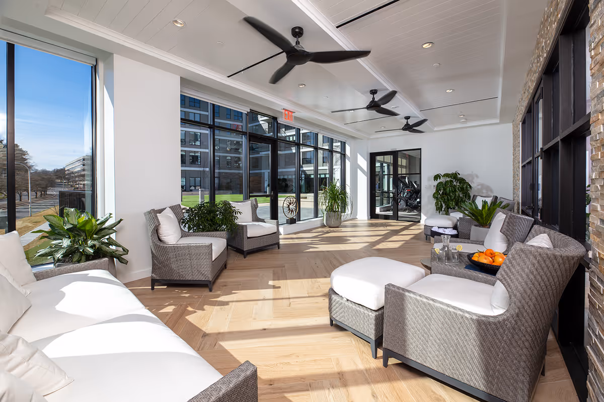 Bright and spacious sunroom with large floor-to-ceiling windows allowing natural light to flood in. The room is furnished with cushioned wicker chairs, sofas, and ottomans arranged around small tables. Several green plants add a touch of nature, and ceiling fans are mounted on the white paneled ceiling. Outside, a grassy courtyard and modern building are visible through the windows.