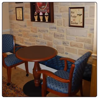 Small seating area with a round table and two upholstered chairs in front of a stone-faced wall decorated with framed ice cream and Coca-Cola signs.