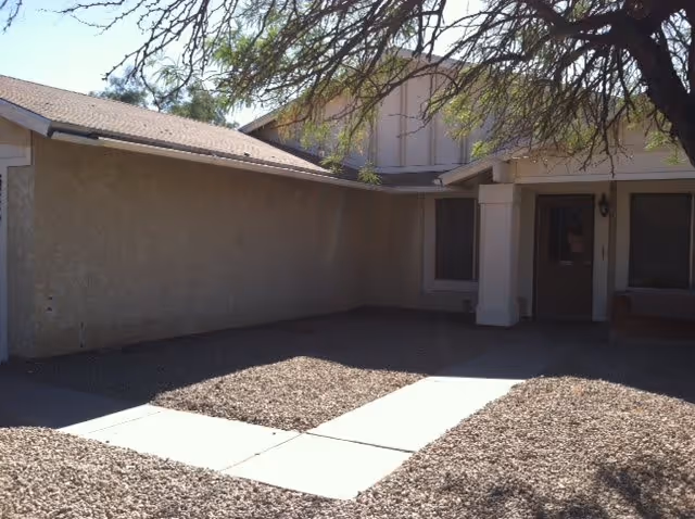 Exterior view of a building with a beige stucco wall and a covered entrance. There is a concrete walkway leading to the door, surrounded by gravel landscaping. A tree with sparse branches is visible in the foreground.