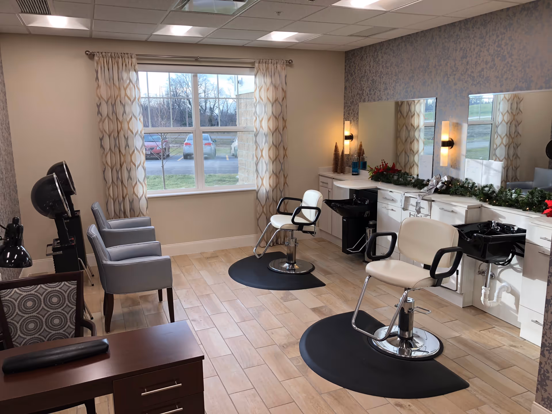Interior view of a salon area in a senior living facility with two salon chairs in front of large mirrors and black wash basins. There are two gray armchairs near a window with patterned curtains, and a wooden desk with a lamp in the foreground. The room has light wood flooring and soft lighting.