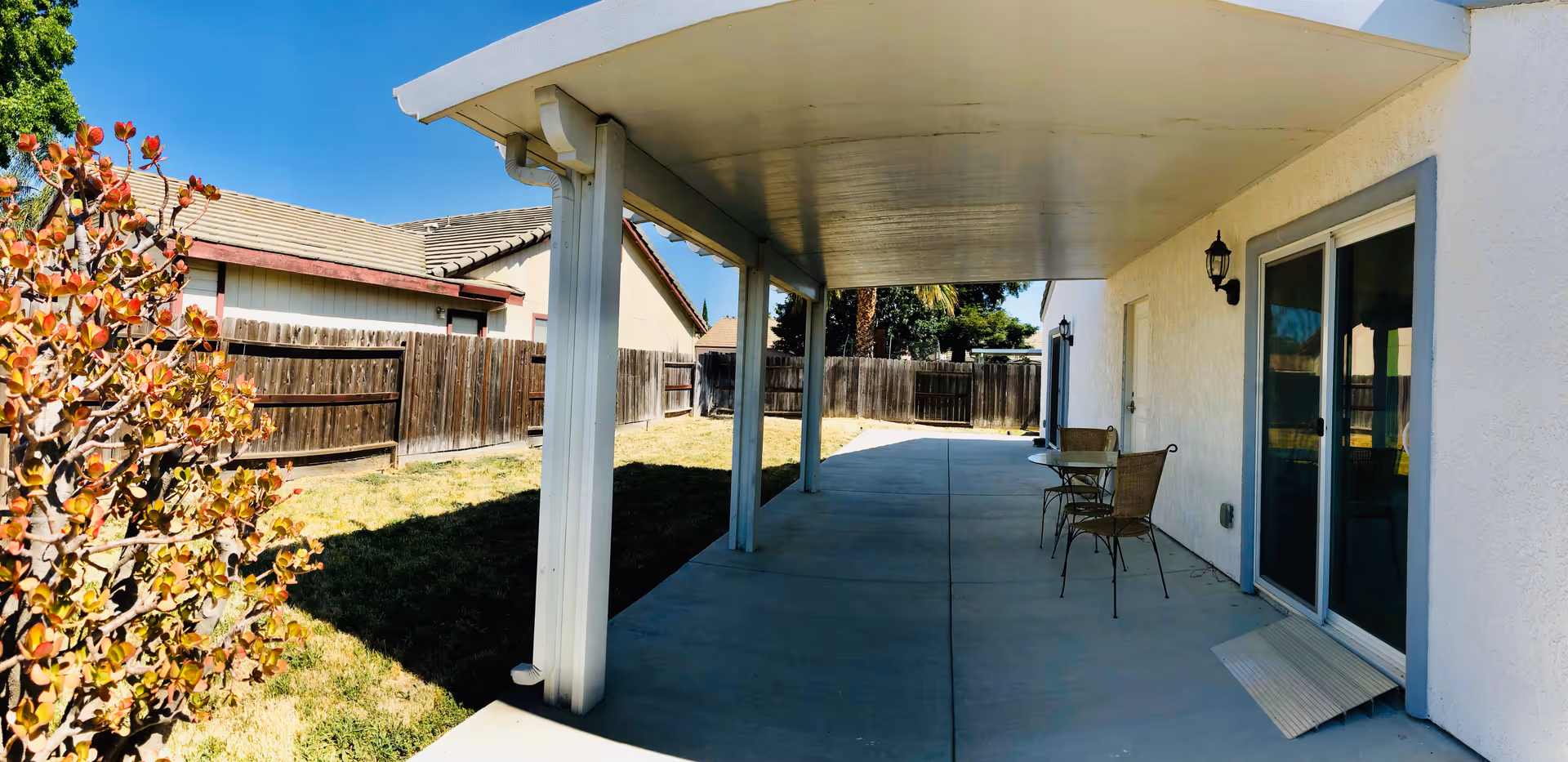 A covered patio area with a concrete floor attached to a white building. There are two metal chairs and a small round table under the patio cover. The patio overlooks a fenced backyard with dry grass and a neighboring house visible. The sky is clear and blue.